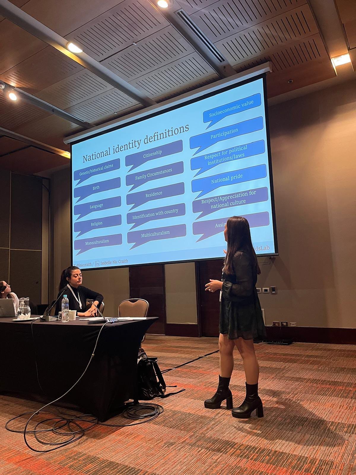Isabelle is looking towards a powerpoint presentation depicting national identity definitions, including birth, language, religion, citizenship and national pride. 