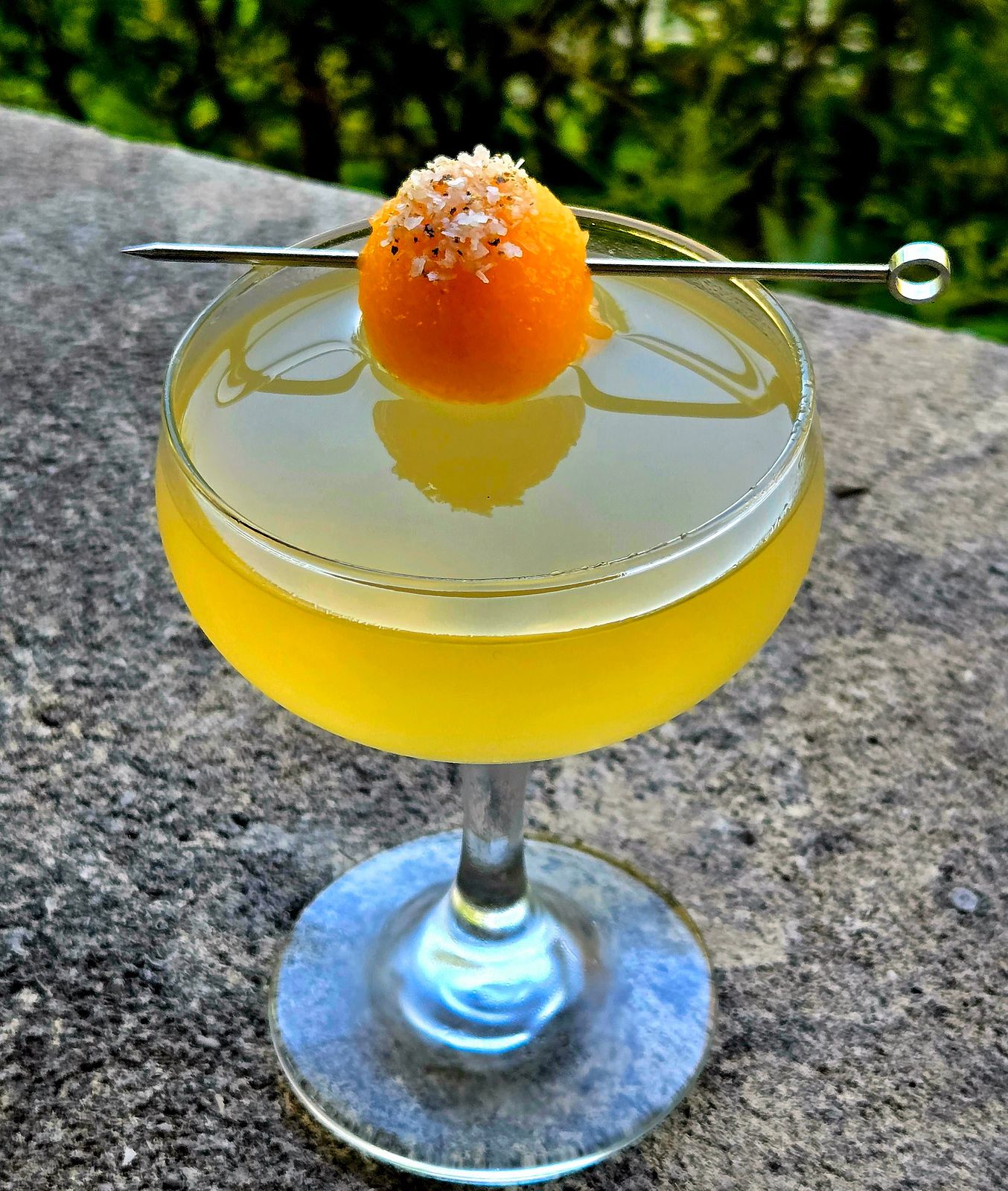 Cantaloma - a cocktail of tequila, cantaloupe liqueur, and pink peppercorn liqueur in a coupe glass sitting atop a stone outdoors with a salt and pepper cantaloupe ball as garnish.