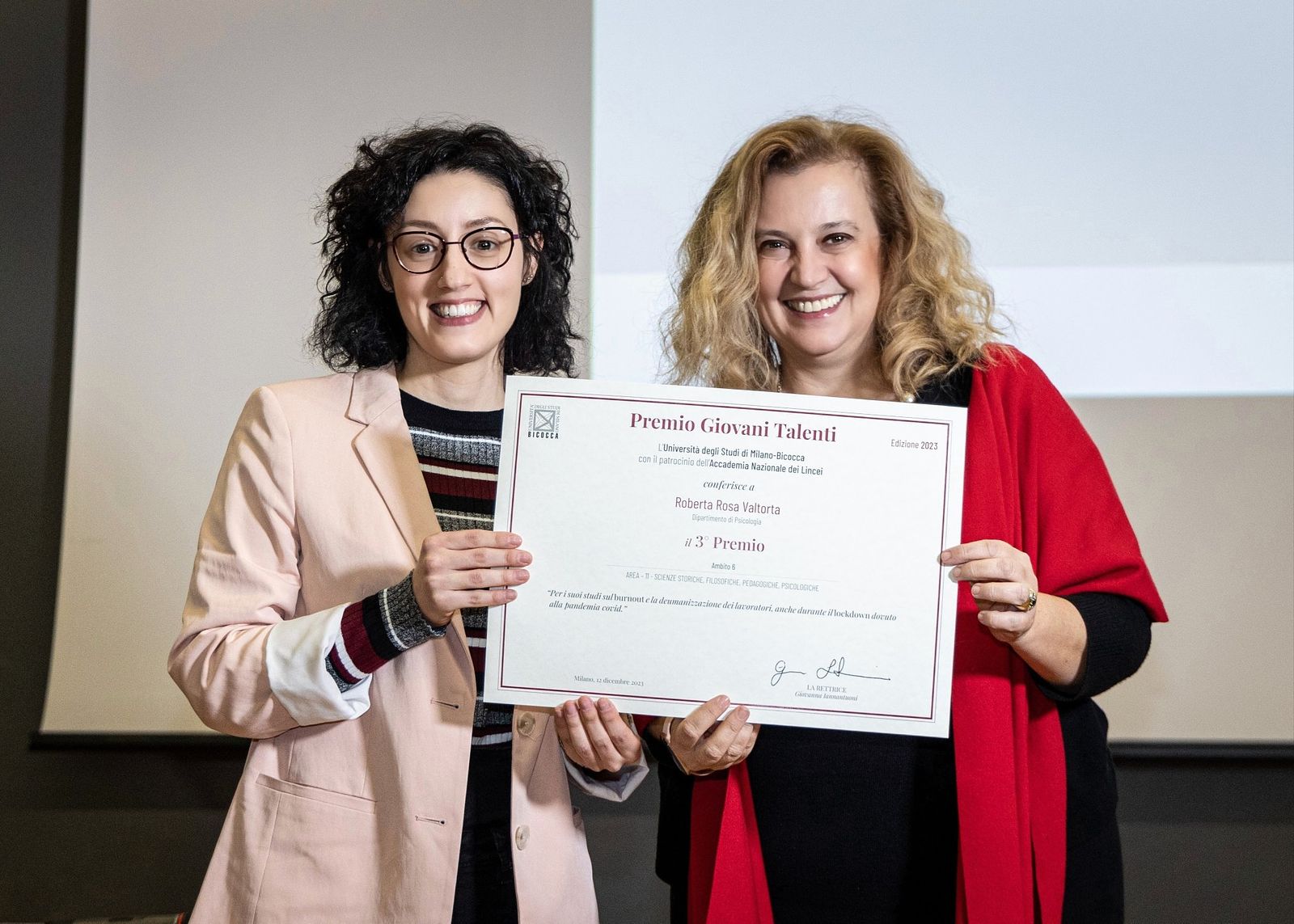 Official award photo: I am wearing a pink jacket and dark shirt, receiving the "Young Talents 2023" award certificate from Giovanna Iannantuoni, then rector of the University of Milano-Bicocca, who is dressed in black with a red shawl.