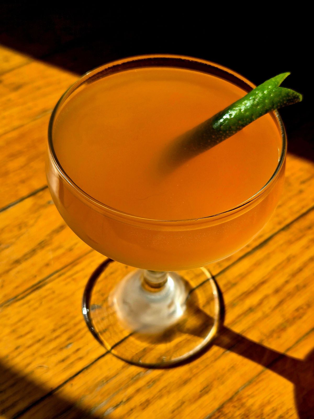 A Pegu Club cocktail in a couple glass with lime lapel garnish sitting on wood in the sun.
