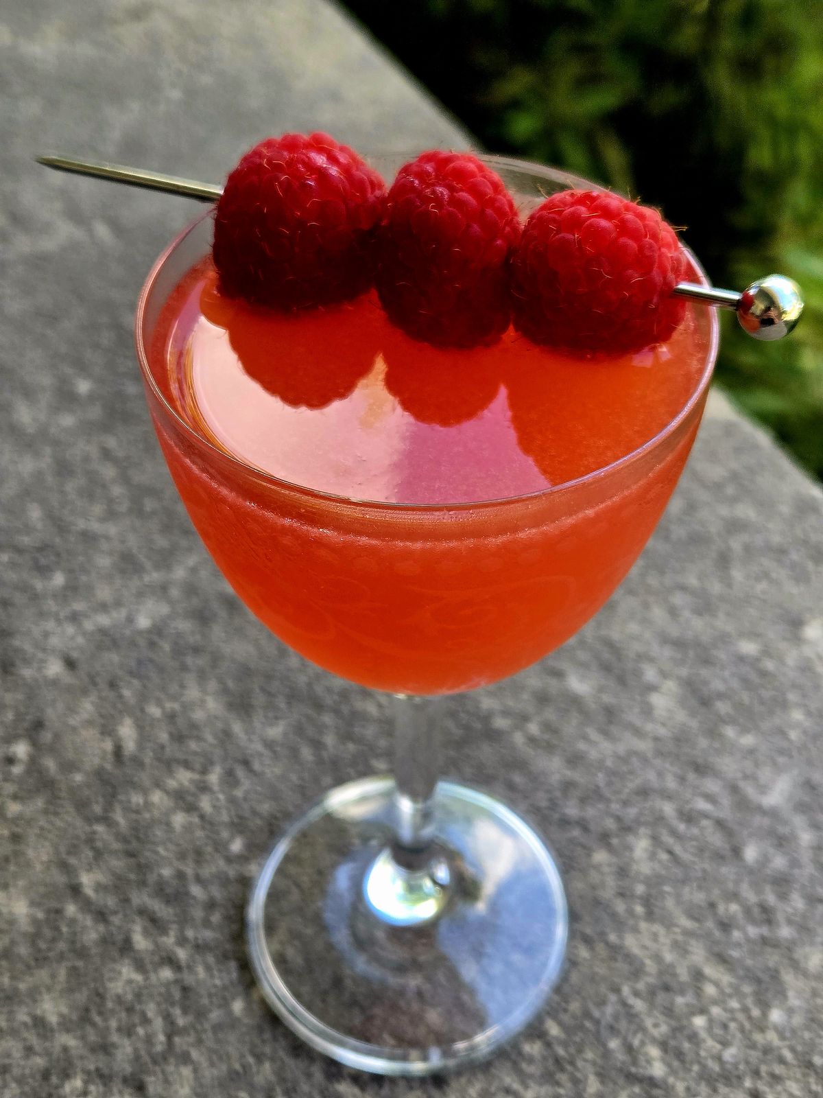 White Lion rum cocktail with raspberry garnish in a Nick and Nora glass atop a stone wall.
