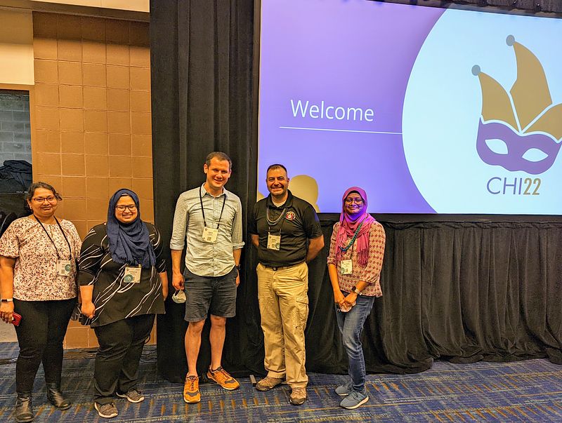 Isha, Tamanna, Jason, Josh, and Sinthia posing in front of the projector screen in the convention center at CHI 2022 in New Orleans