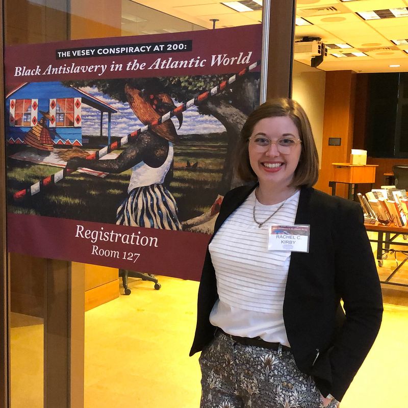 Rachel Kirby, a white woman with short light brown hair, stands in front of a poster that reads "The Vesey Conspiracy at 200: Black Antislavery in the Atlantic World." The poster also shows a painting of a man and woman tending to a rice crop in front of a blue house. The painting is by artist Jonathan Green.