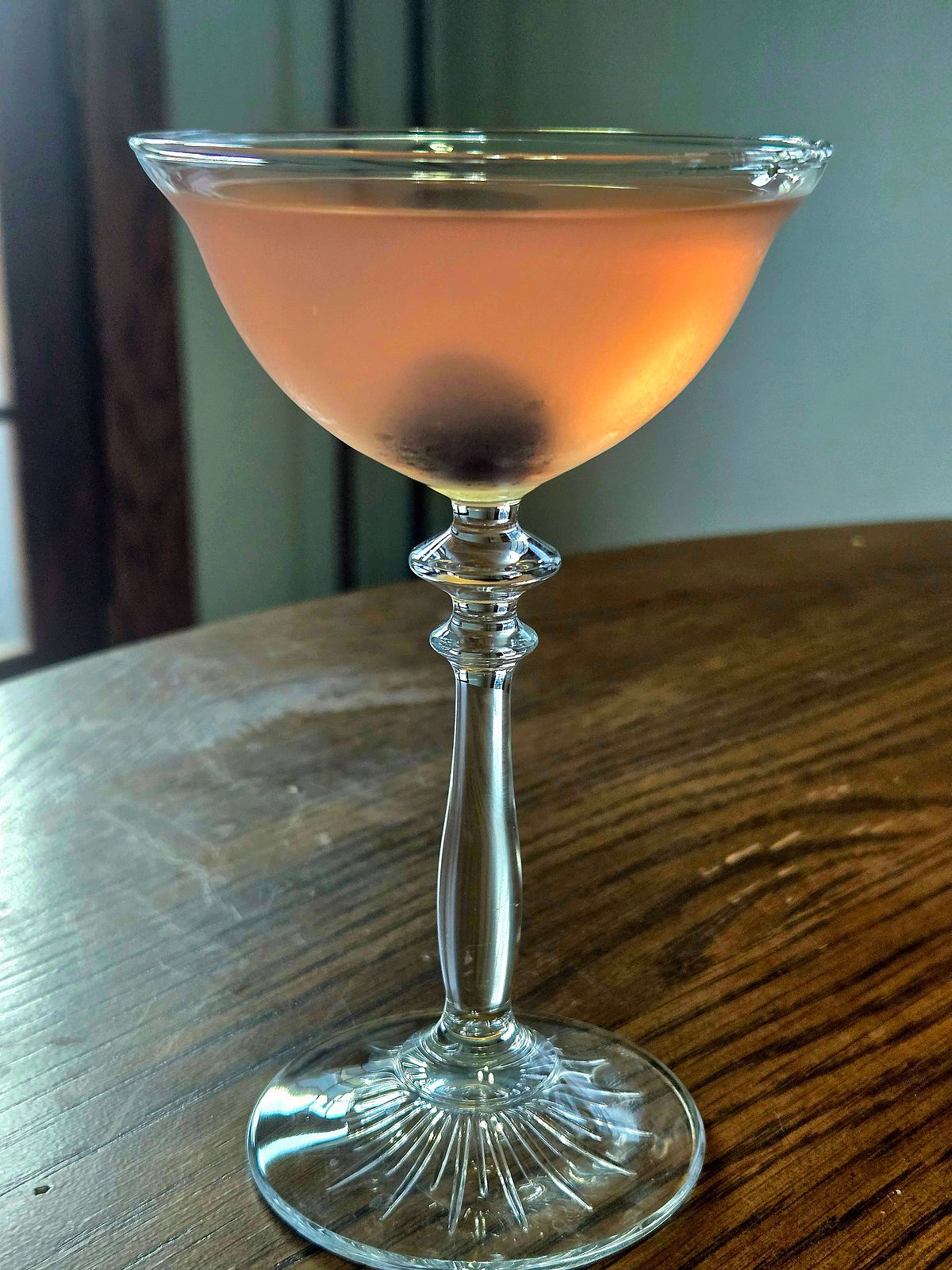 A monkey gland cocktail in a coupe glass with cherry garnish sitting atop a wooden table in profile view.