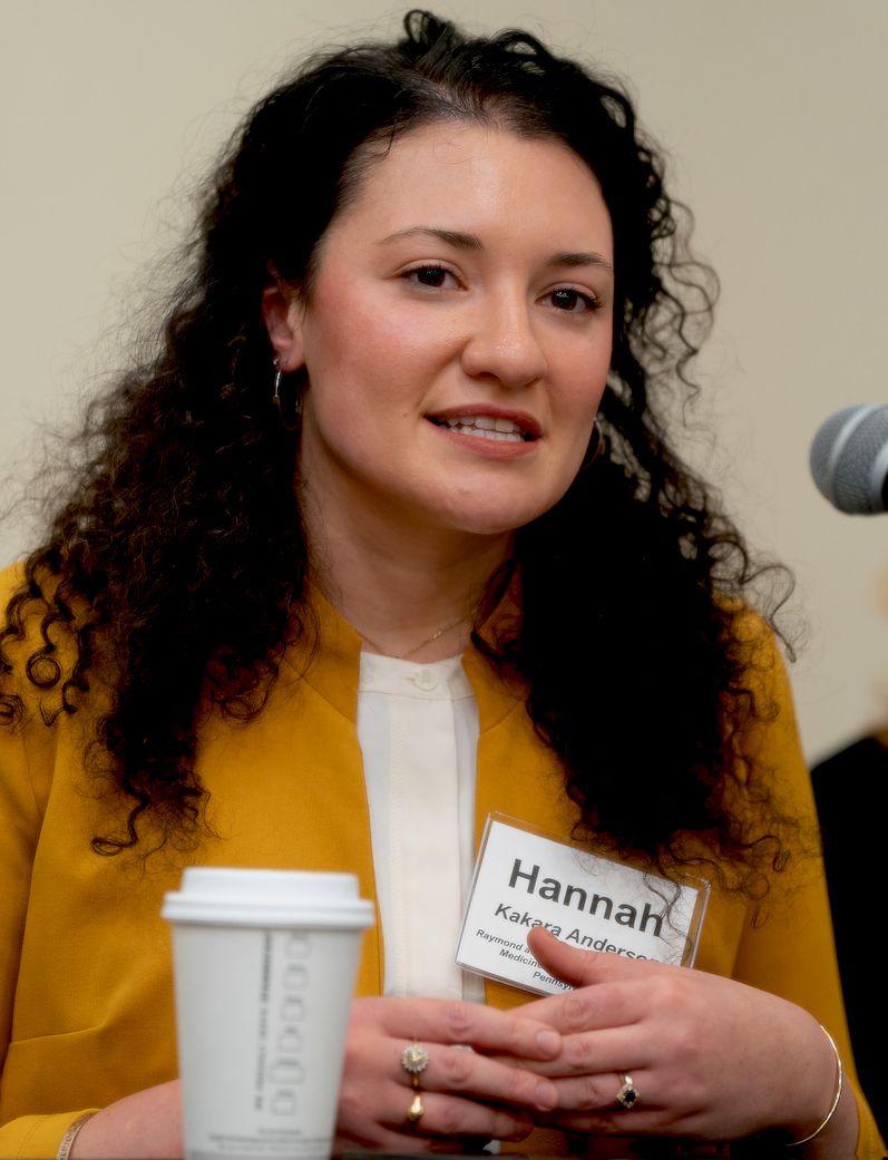 Hannah is sitting at a table with a microphone in front of her. She is talking and look off camera. She is wearing a yellow blazer and a white shirt. She has light skin and black curly hair that comes below her shoulders.