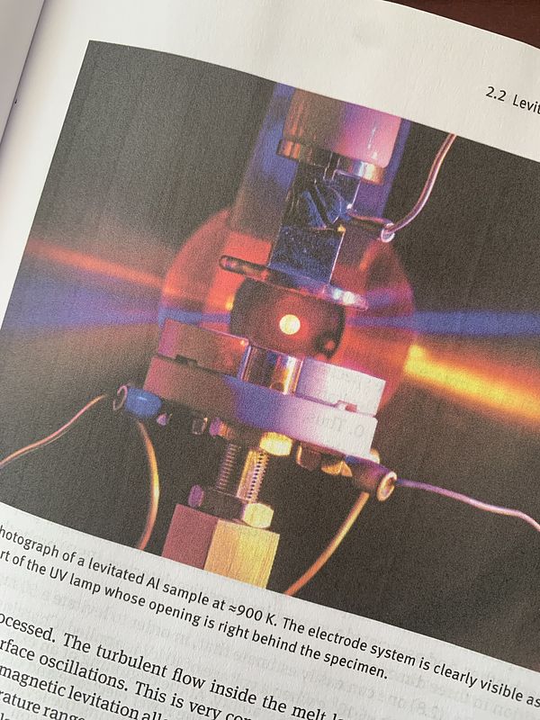 Four gray electrodes are surrounding a yellow/orange glowing and molten metal sample, which is levitating in an electrostatic field.