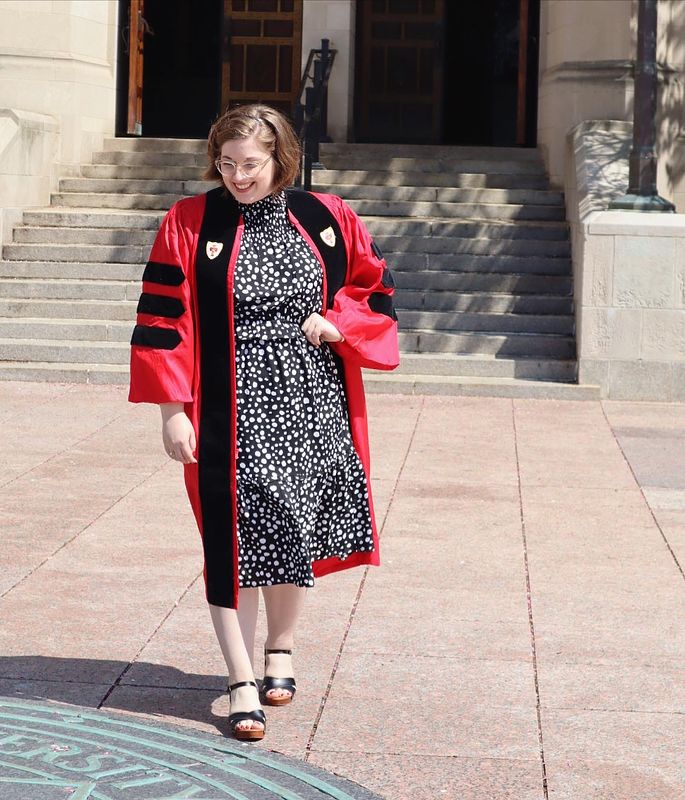 Picture of a white woman (Rachel Kirby) in a red doctoral robe and black and white dress looking down.