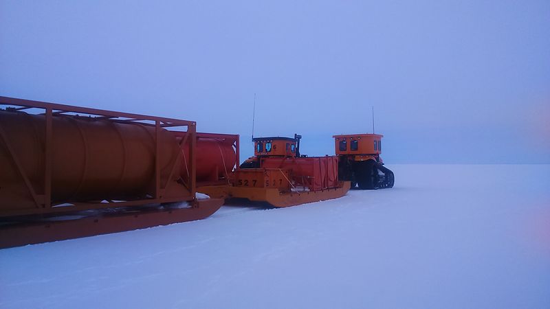 Large orange catepillar tractors with rubber tracks pulling sleds of equipment and fuel tanks on Antarctic ice.