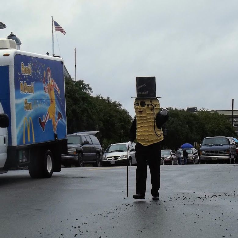 A figure walks down a street wearing a top top, a peanut-shaped costume covering their face and torso, and holding a cane - they are dressed like "Mr. Peanut." To their right is a truck featuring a McDonald's advertisement with Ronald McDonald.