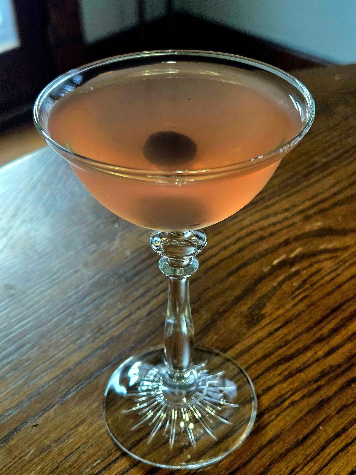 A monkey gland cocktail in a coupe glass with cherry garnish sitting atop a wooden table.