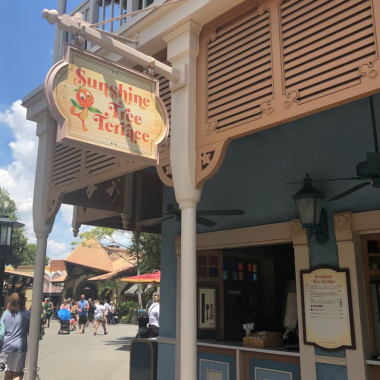 A sign in front of a snack stand window reads "Sunshine Tree Terrace" in orange text and shows the "Orange Bird" - a character with a citrus orange for a head and green leaves for wings.