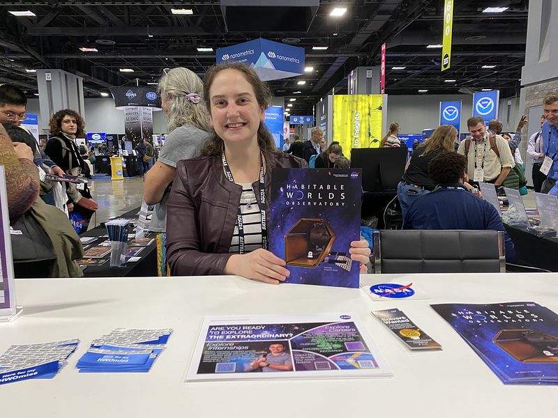 Jessica Noviello sits at a table holding a flyer for the Habitable Worlds Observatory. There are many people and signs in the background behind her. In front of her on the table are NASA stickers and more flyers advertising NASA science activities. 