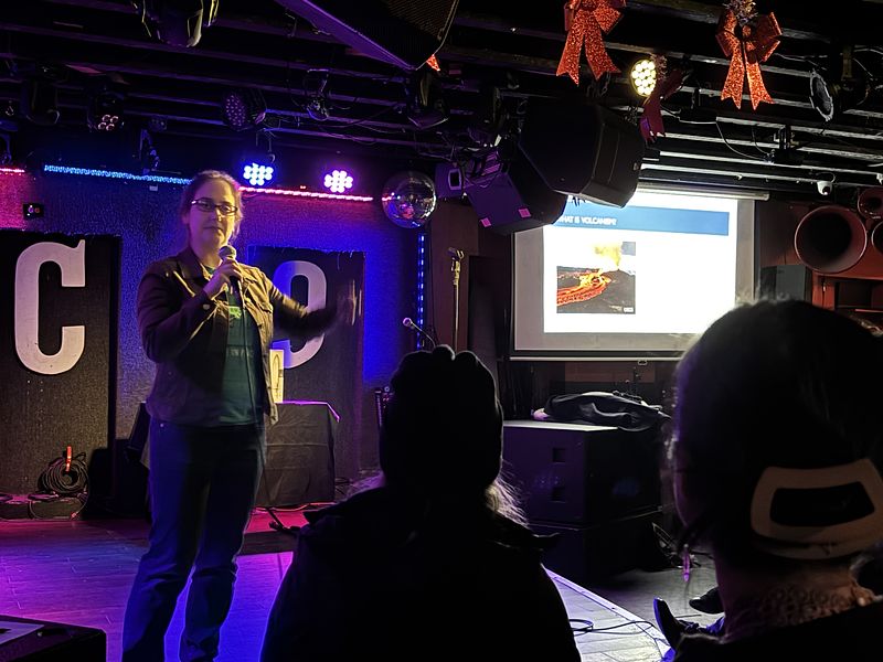 Jessica is standing on a stage, holding a microphone. Her left hand is pointing to a screen in the background with a picture of a lava eruption on it. 