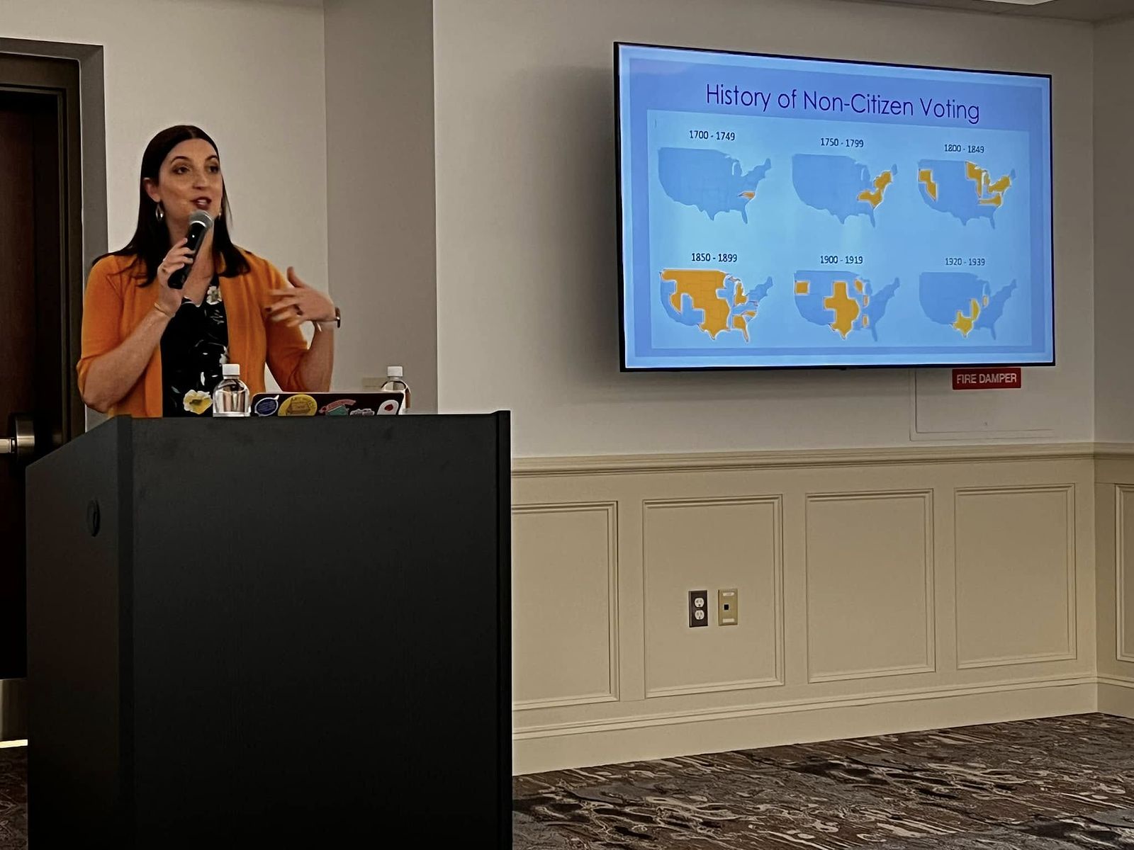 Dr. Gunter in a blue dress with white and yellow flowers and a yellow blazer in speaking into a microphone in front of a slide showing the History of Non-Citizen Voting in maps