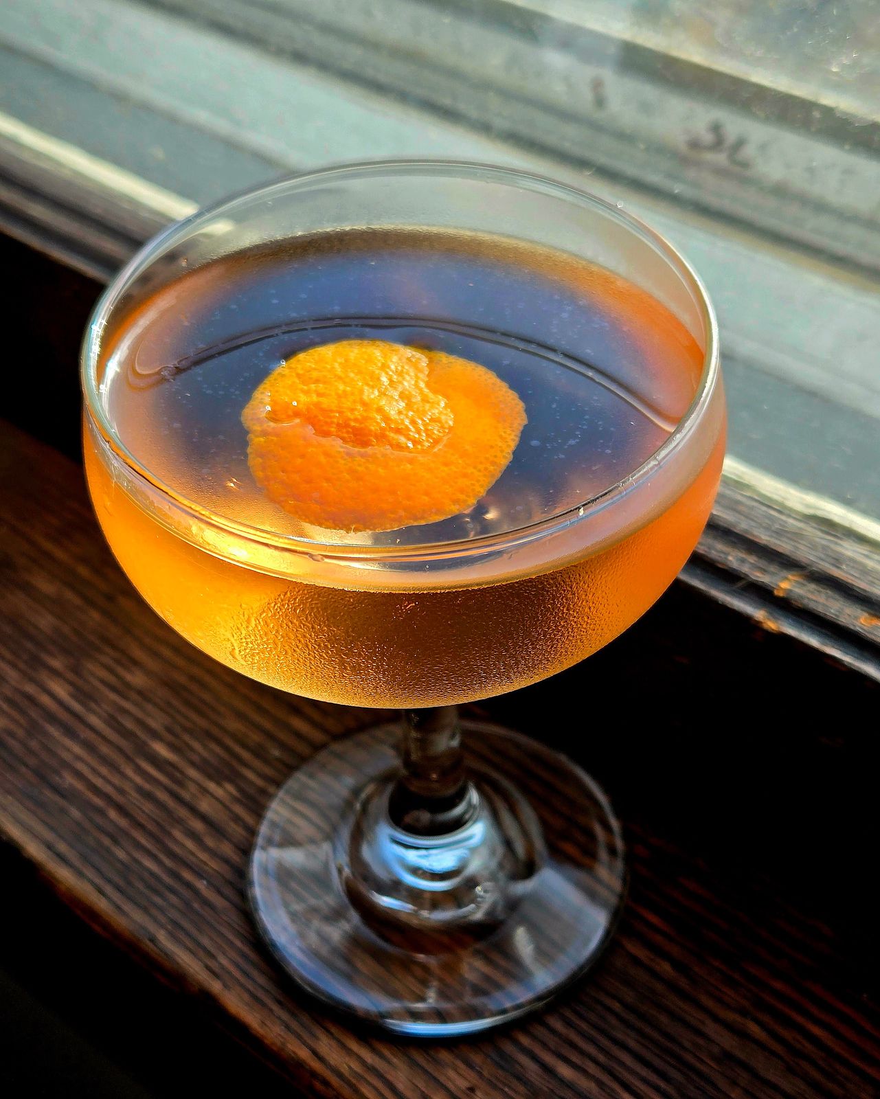 A Presidente cocktail with orange island garnish in a coupe glass sitting in a window seal.