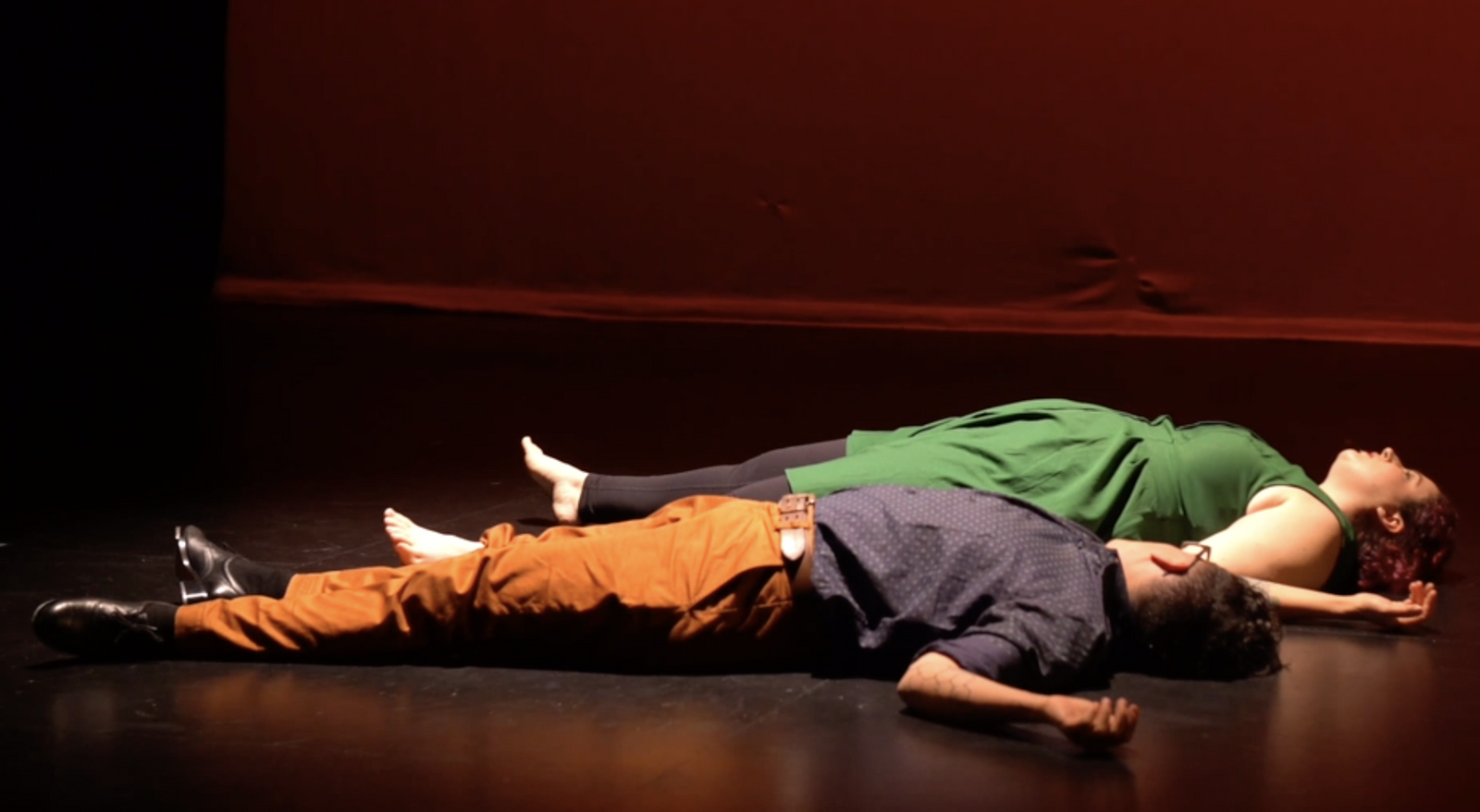 A still image of two dancers lying on a black stage in front of a red scrim