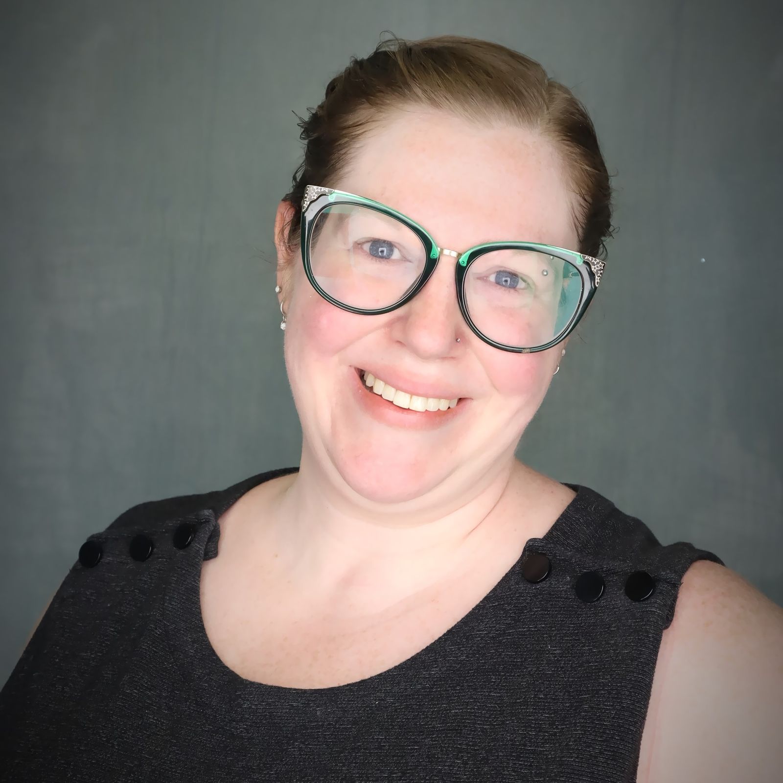 Maggie Albro smiles into the camera. She is a white woman with light brown hair worn in a low bun. She is wearing large green cat-eye style glasses and a dark gray dress with buttons across the shoulders.