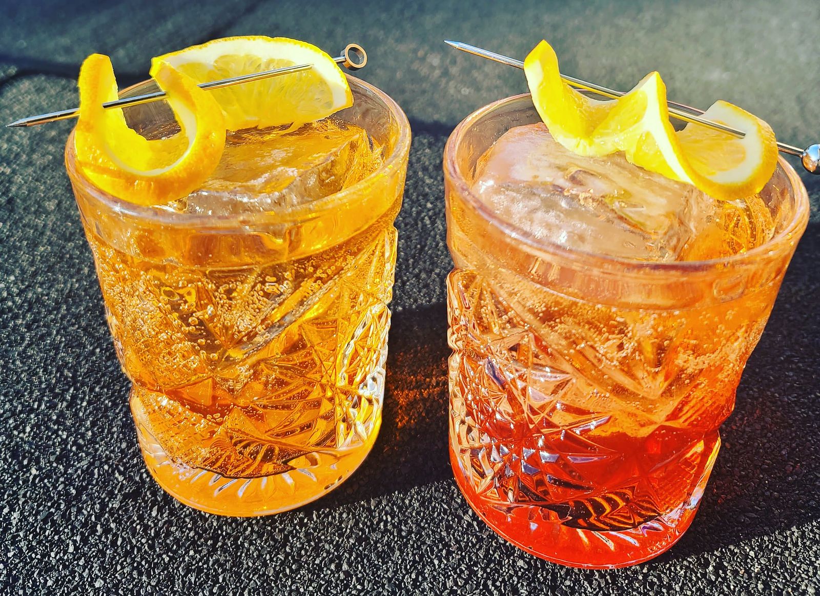 A Contessa and Negroni cocktail, side by side, with citrus garnish on a roof in the sun.