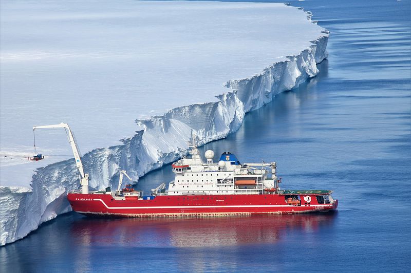 Red  & white polar research ship pushing against towering Antarctic shelf and offloading equipment onto ice shelf with lare crane at front of ship