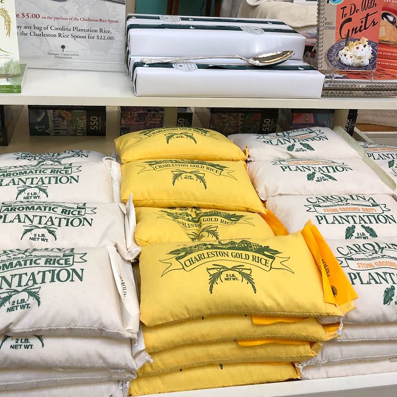 Fabric bags of rice are stacked on a display shelf. The bags on the right and left are white with green font, and the bags in the center are yellow with green font. The middle bags are of "Charleston Gold Rice."