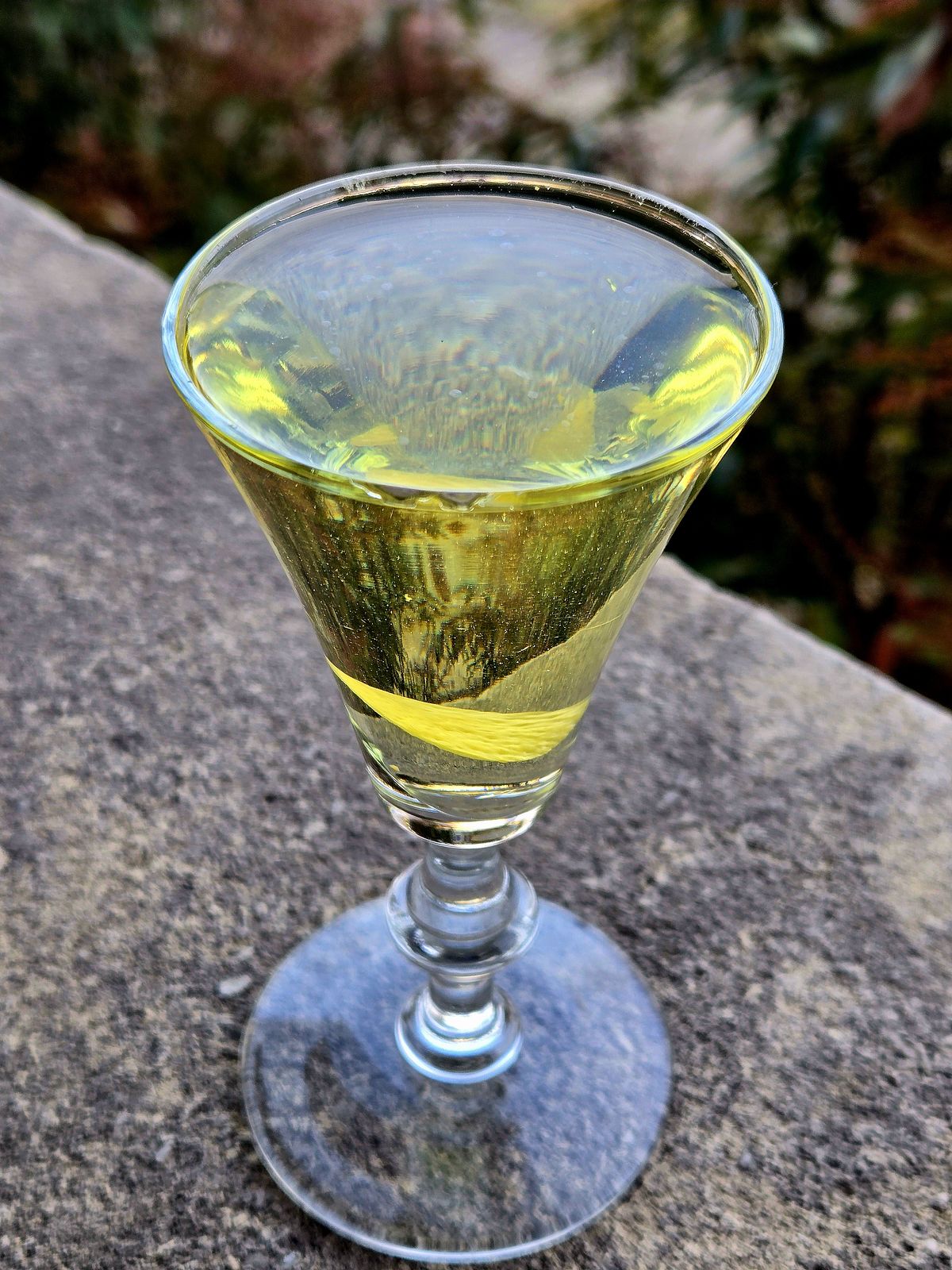 Alaska cocktail with lemon disc garnish in a sherry glass sitting atop a stone wall.