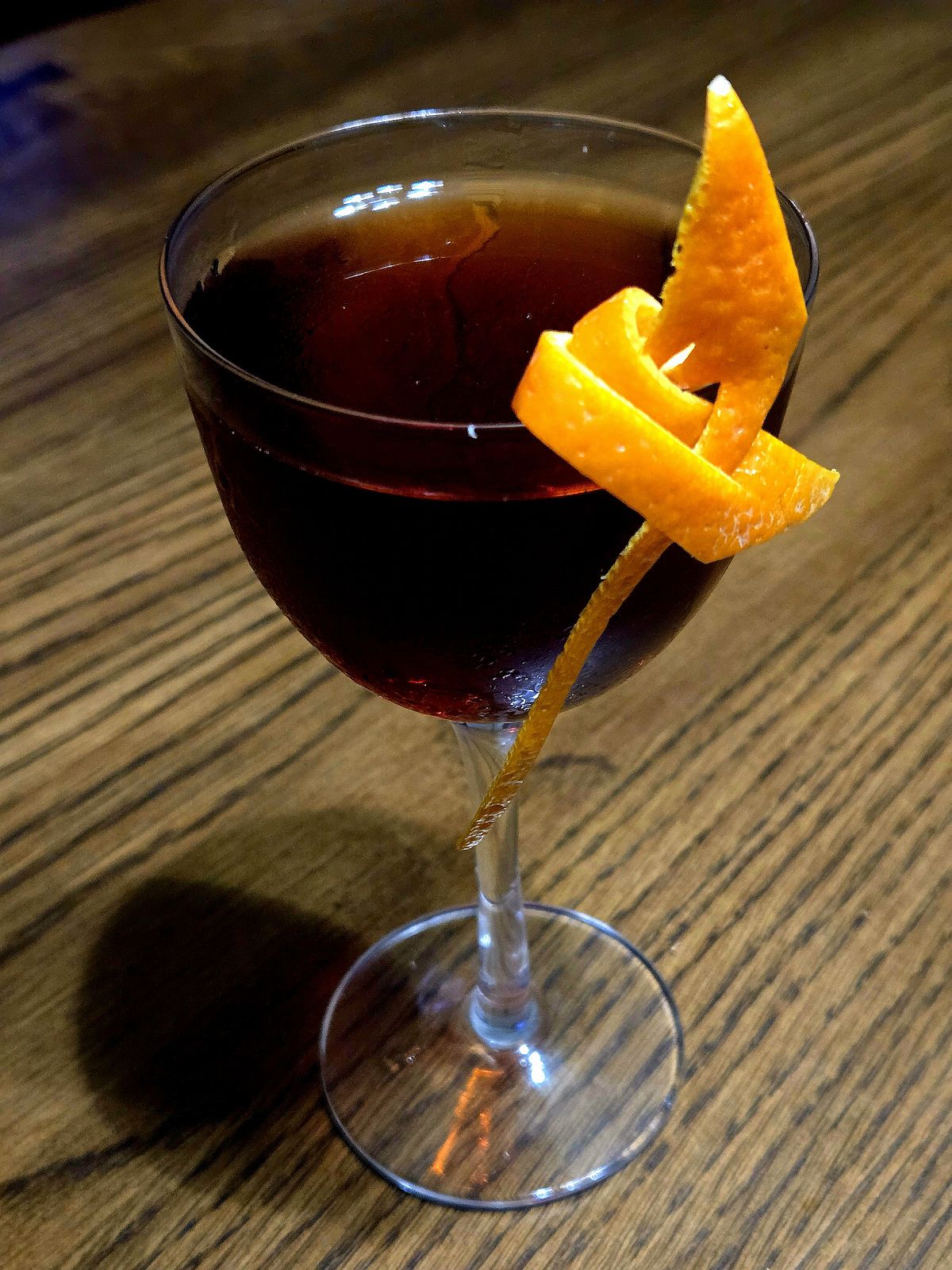 Nice One cocktail in a Nick and Nora glass sitting atop a wooden table with orange twist garnish.