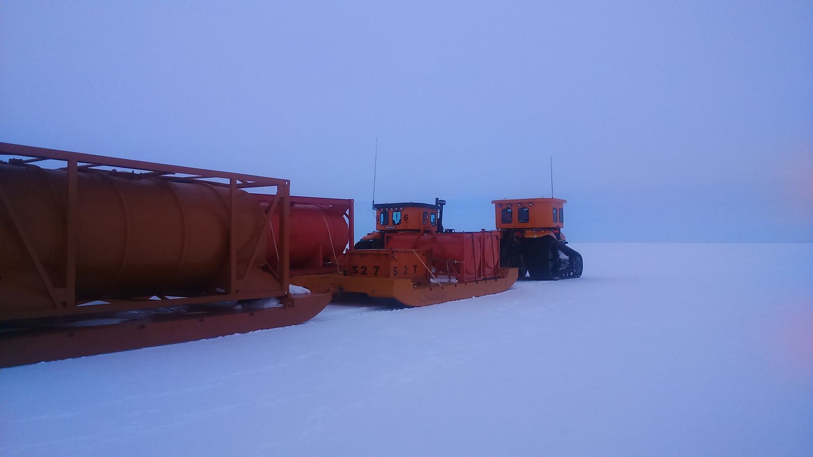 Large orange catepillar tractors with rubber tracks pulling sleds of equipment and fuel tanks on Antarctic ice.