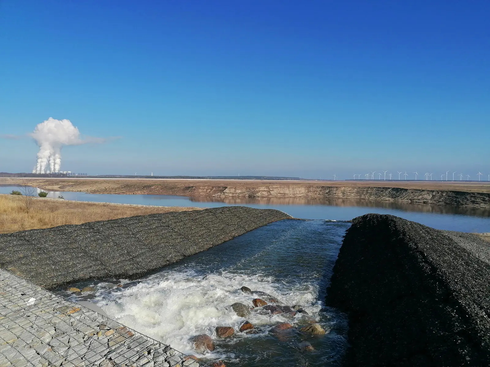 Germany, Brandenburg, Cottbus (2021): Flooding of an end-of-life open pit lignite mine to create an artificial lake, with both a coal power plant and a wind farm in the background