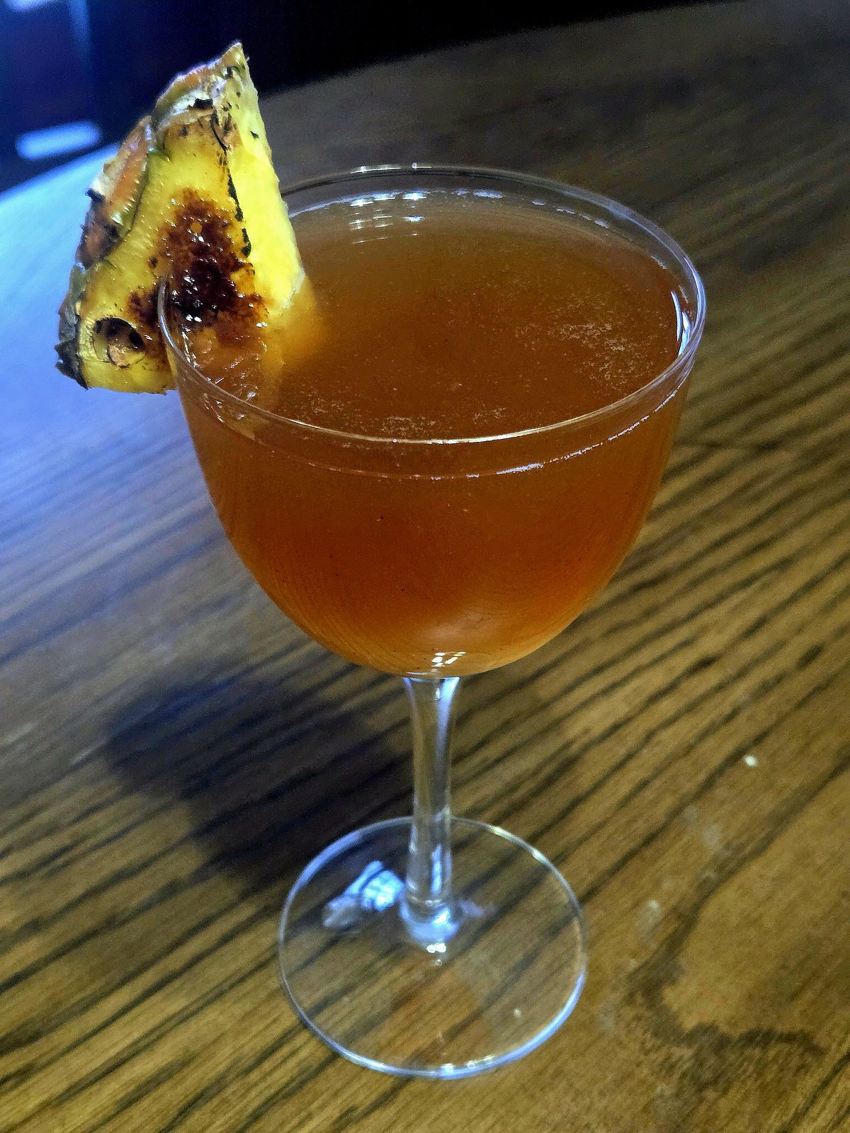 A cocktail, Saint Cecilia's Society Punch, in a Nick and Nora glass sitting atop a wooden table with pineapple wedge garnish.