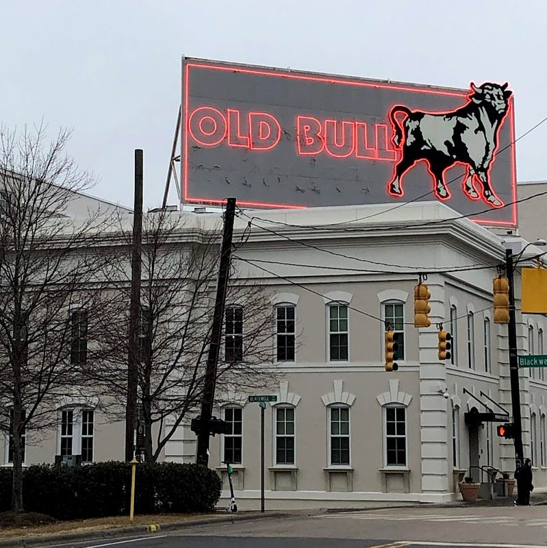 A gray billboard sits upon a beige and white Italianate building. The sign has a black and white graphic of a bull on the right, and has red neon lighting that reads "OLD BULL"