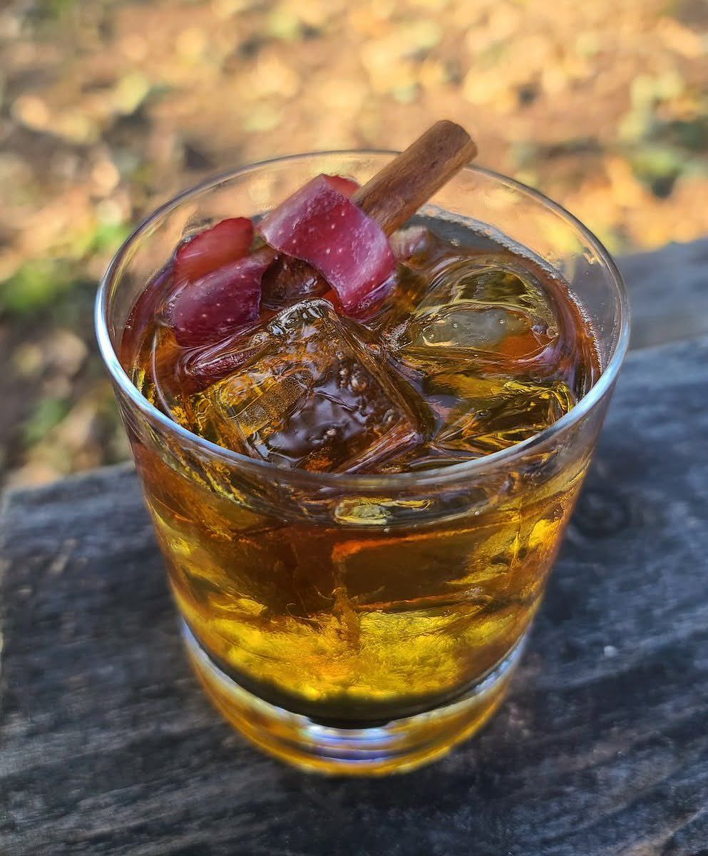 "Old EP" - An Apple brandy cocktail in a rocks glass with cinnamon stick and apple twist garnish atop a wooden picnic table.