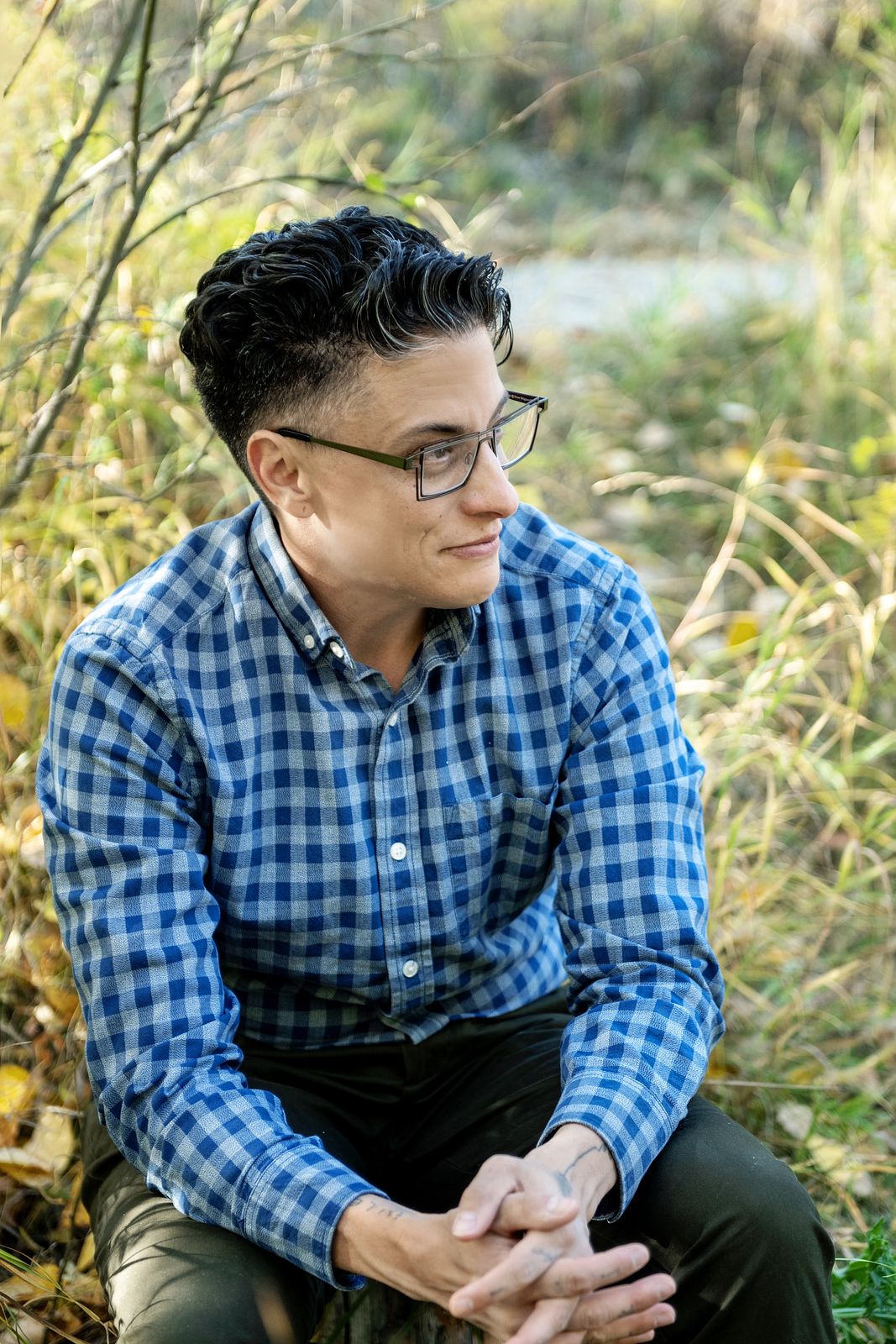 An image of me, Nathan, sitting among grasses and tree branches. I have light brown skin, dark brown and grey short-cut hair, and visible tattoos on my hands and wrists. I am wearing business casual clothes and rectangular glasses.