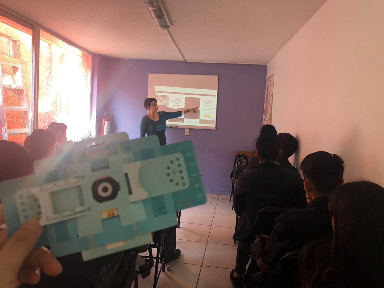 Photograph of a classroom where a woman at the front is pointing to a screen showing the Foldscope website. In the foreground, the photographer has held a Foldscope up to the camera.