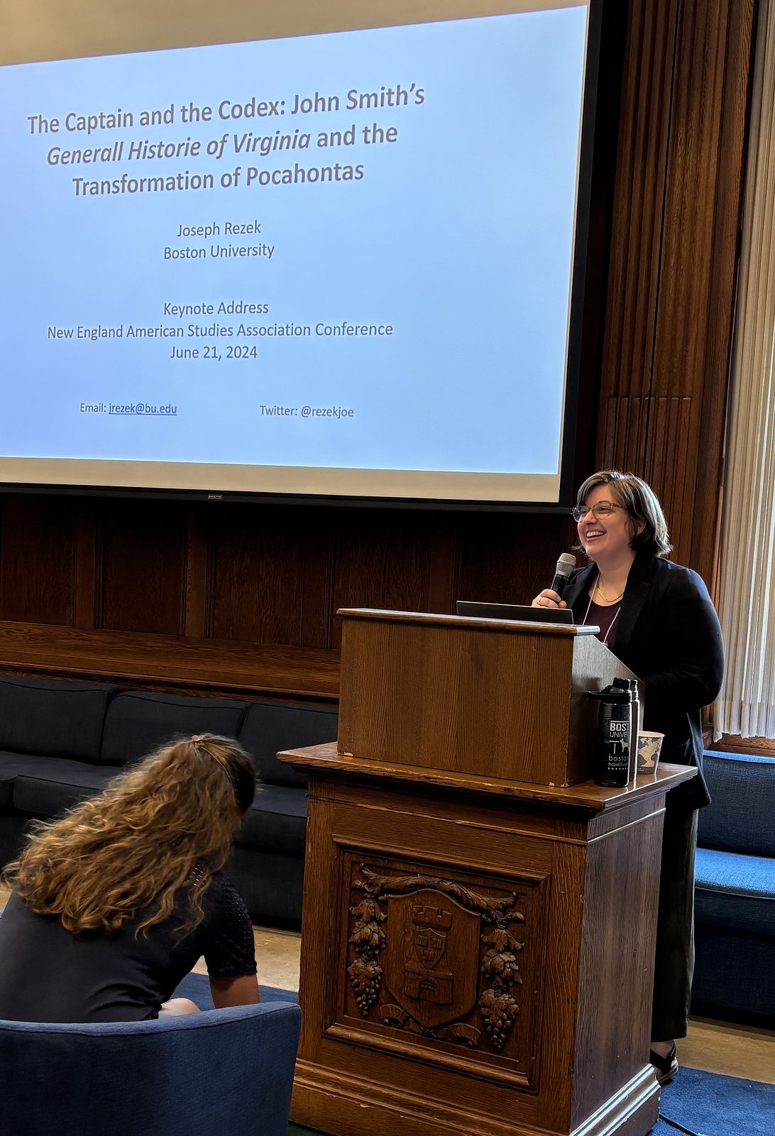 Rachel Kirby, a white person with short brown hair, holds a microphone and stands behind a podium. A screen in the background reads "The Captain and the Codex: John Smith's Generall Historie of Virginia and the Transformation of Pocahontas."