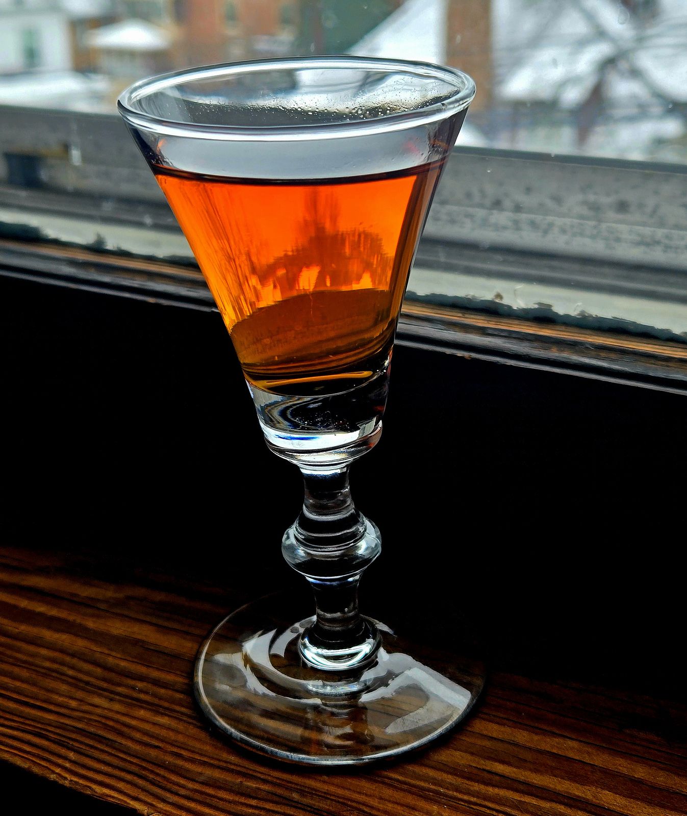 A sherry glass of Medjool Date Sipping Liqueur sitting on a vintage window sill with snow capped houses in the background.