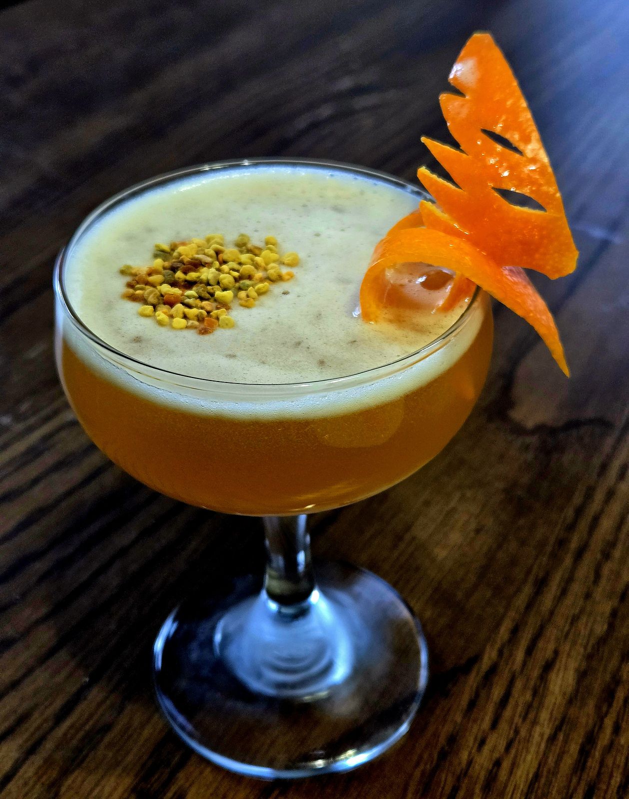 The Birds and the Bees cocktail with bee pollen and orange hummingbird garnish in a coupe glass atop a wooden table.