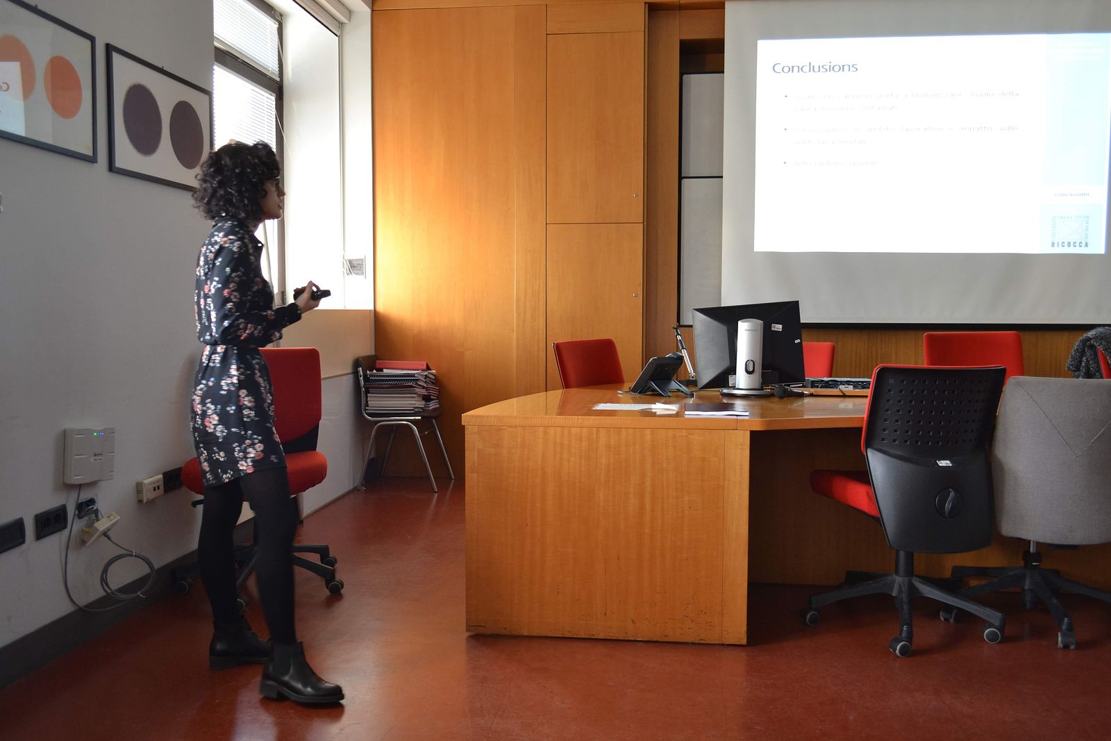I am standing in a classroom at the University of Milano-Bicocca, wearing a black dress, defending my PhD thesis. A slide titled "Conclusions" is visible in the background.