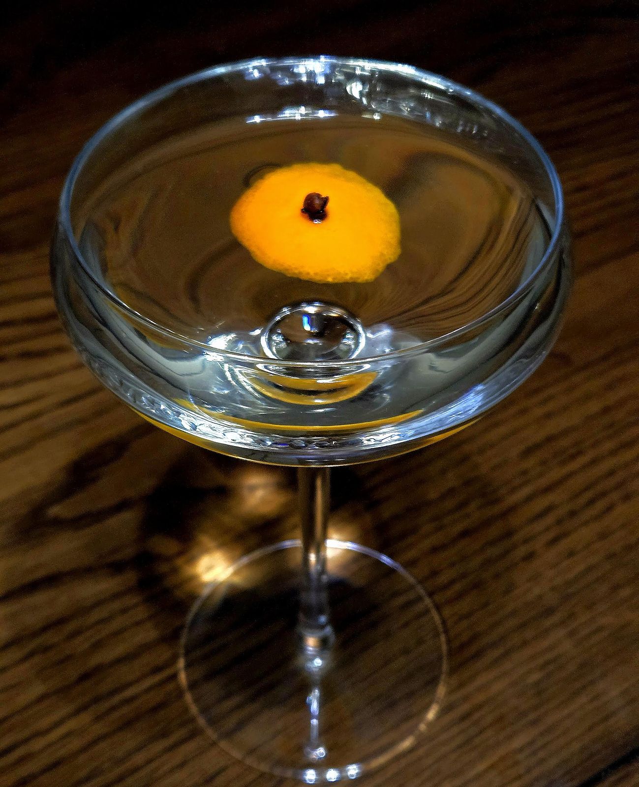 1906 Martini in a Coupe glass with flamed orange disc studded with clove as garnish atop a wooden table.