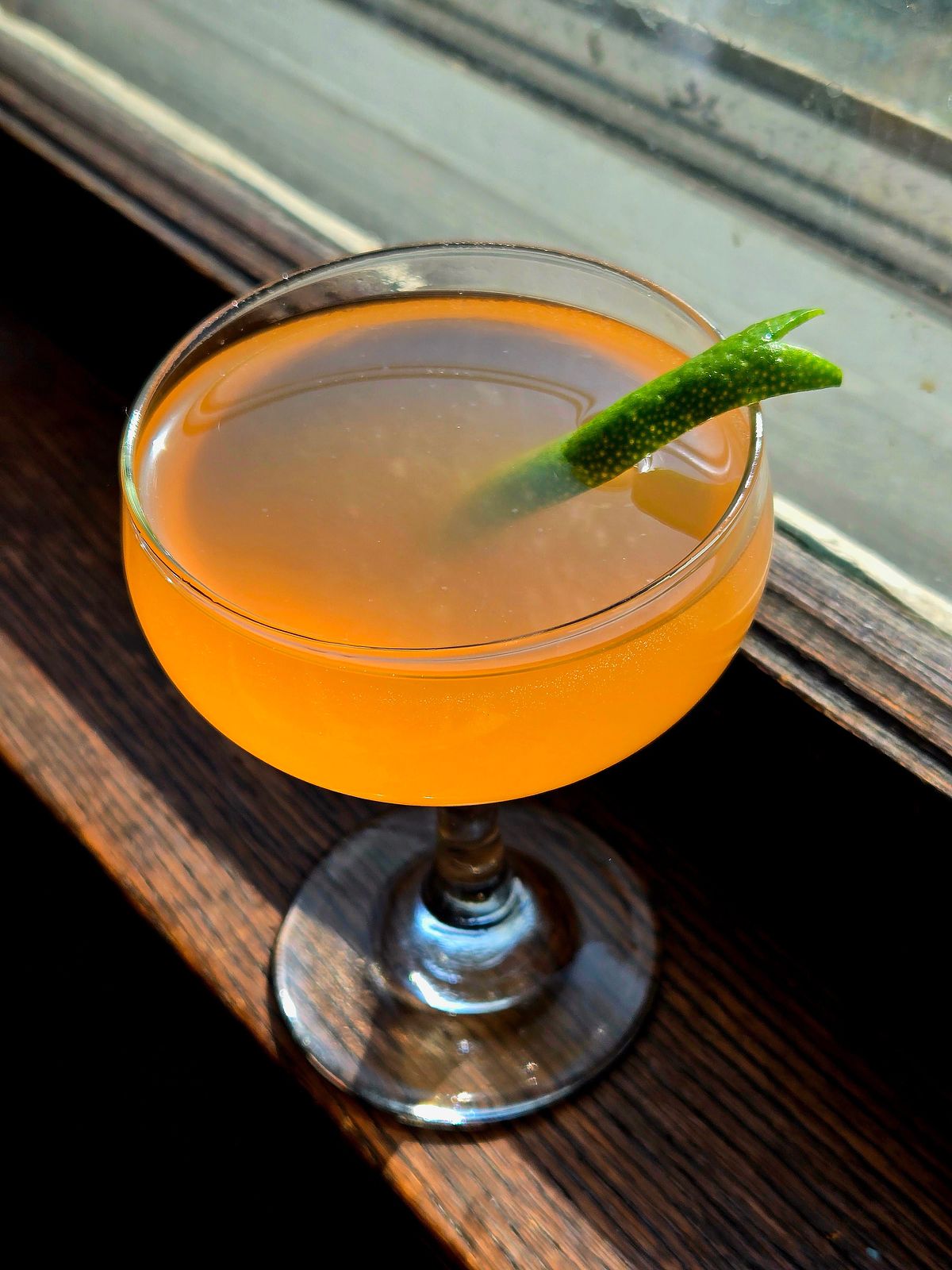 A Pegu Club cocktail in a couple glass with lime lapel garnish sitting on a window seal.