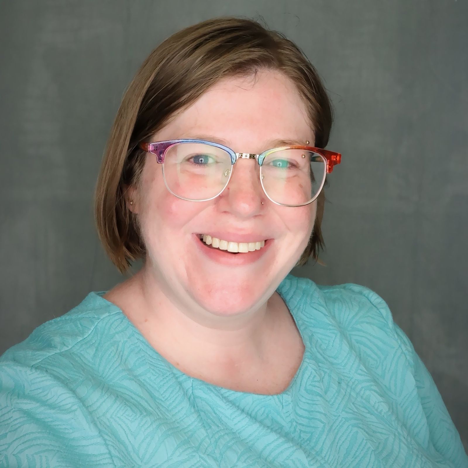 Maggie Albro, a woman with a mousey brown bob, multi-colored glasses, and rosy cheeks, smiles into the camera.