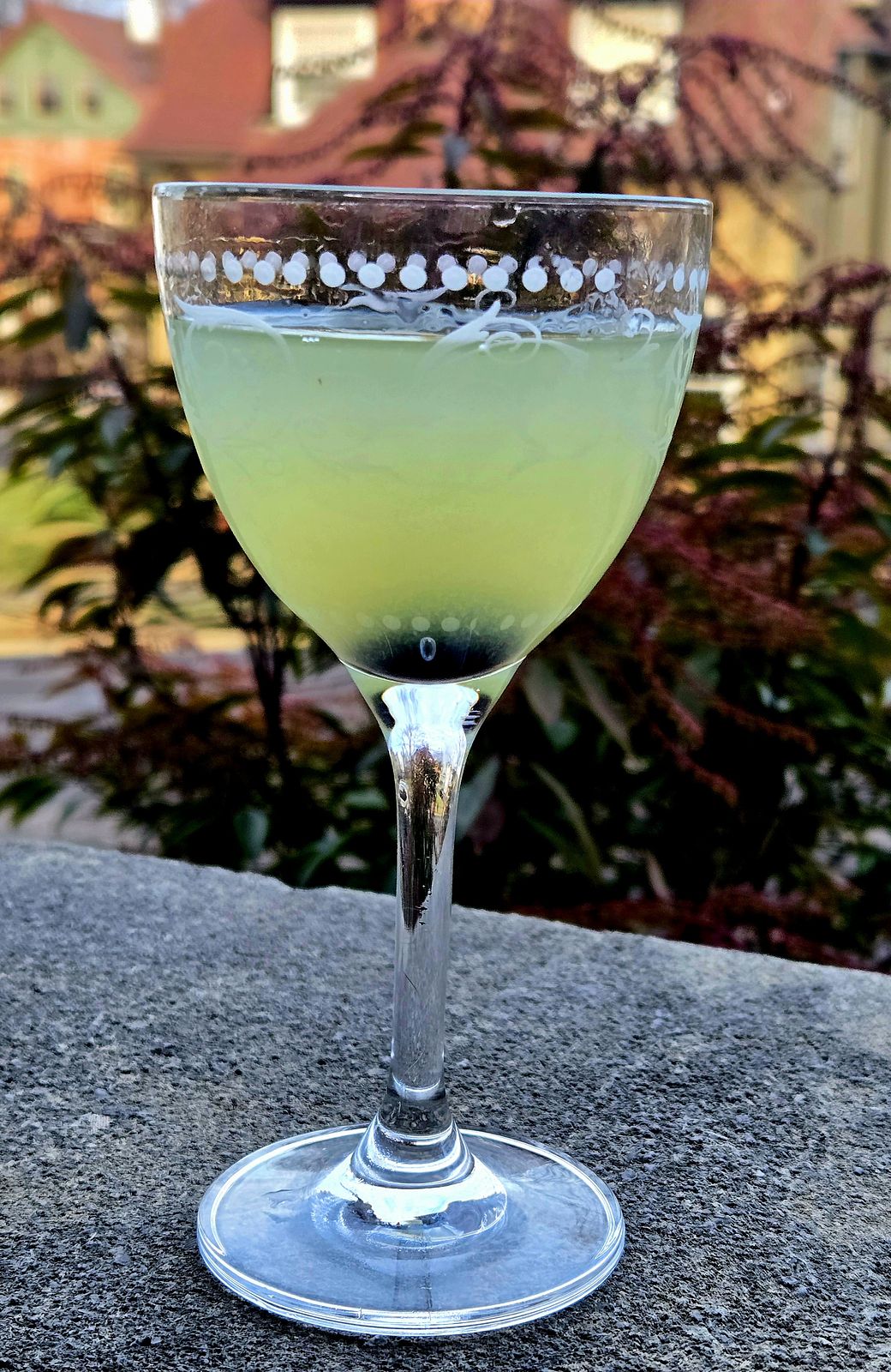 The Last Word cocktail in a Nick and Nora glass with Luxardo Cherry garnish atop a stone wall.