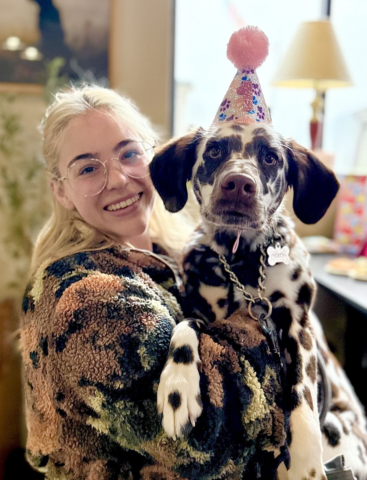 Picture of me smiling while holding my Dalmatian, Pigeon, who looks at the camera and wears a birthday hat.