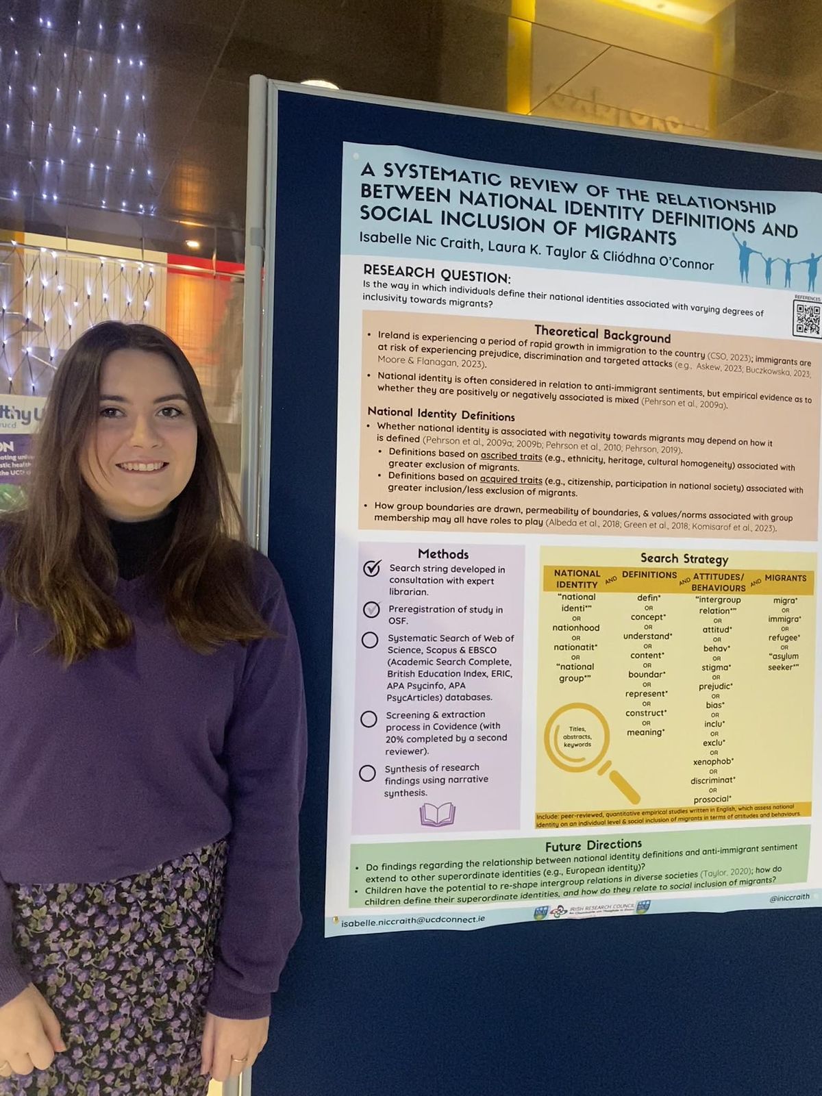 Isabelle presenting her poster at the UCD Psychology Aidan P. Moran Research Day, 2023 - she is pictured smiling beside a research poster entitled "A Systematic Review of the Relationship Between National Identity Definitions and Social Inclusion of Migrants"