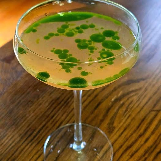 "Byblos" - a cocktail version of a classic watermelon and feta salad having its roots in the levant. It sits atop a wooden table in a coupe glass. Its pink with green "floating" islands of fresh basil oil.
