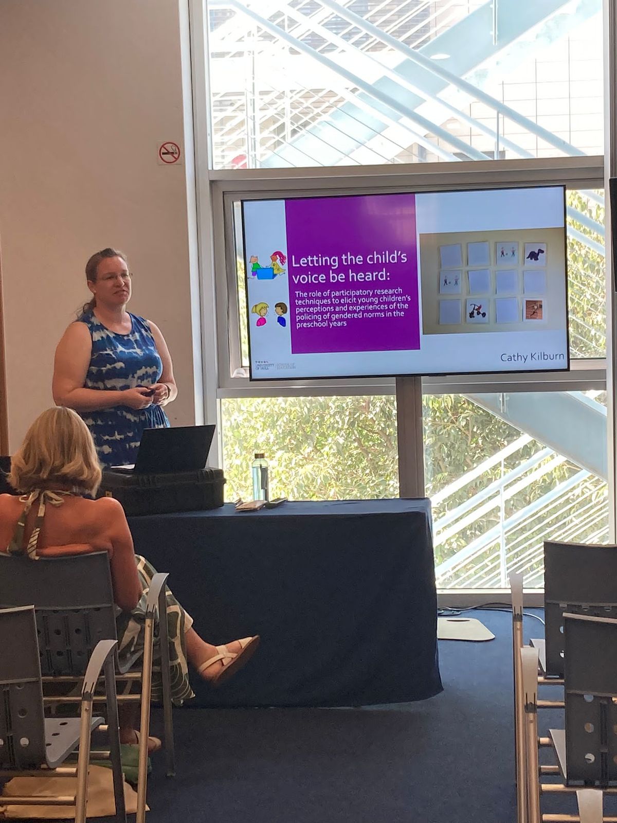 A lady standing at the front of the room next to a large screen presenting to an audience.