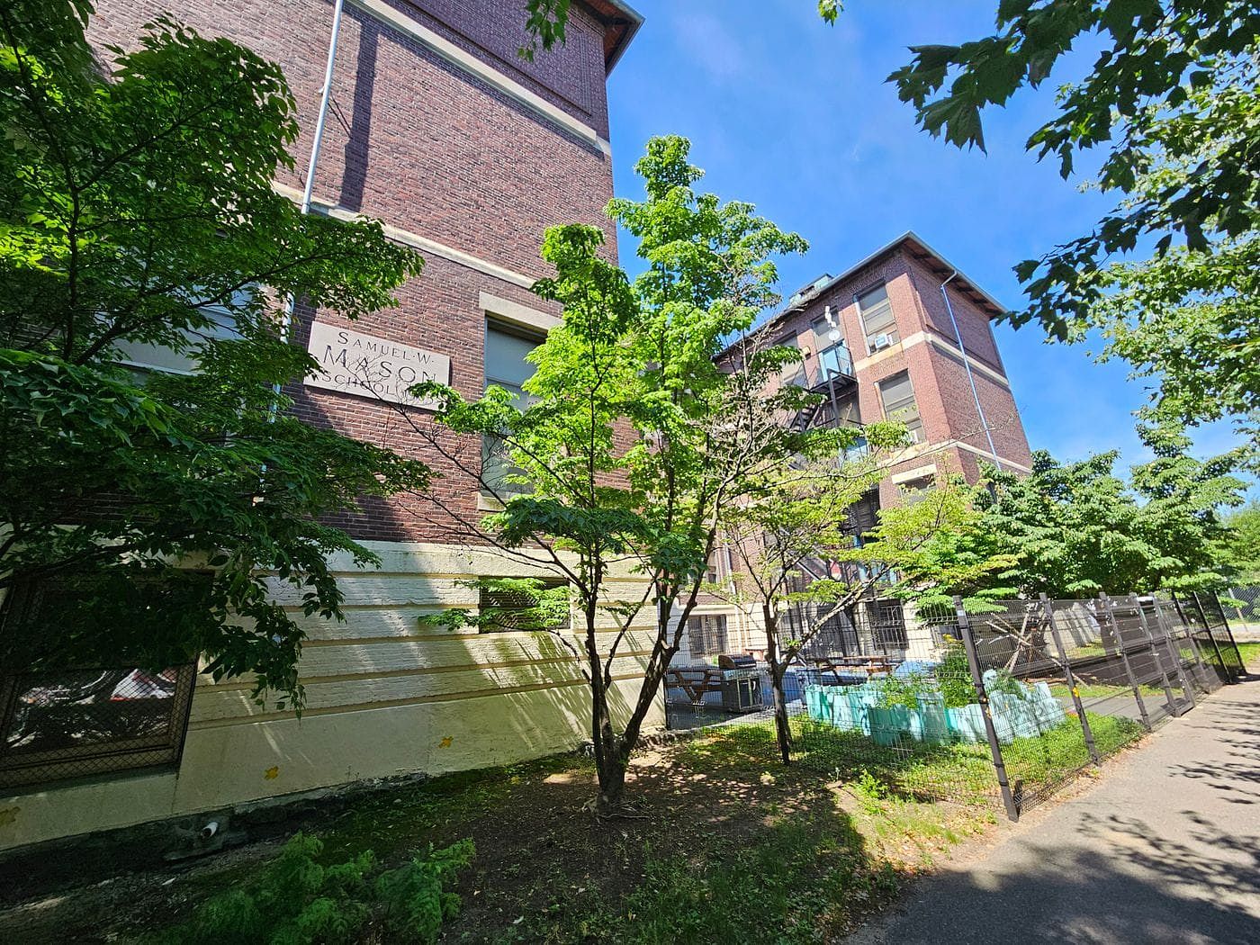 Looking up at a brick building, with a stone sign saying "Samual W. Mason School," just behind green leafy trees