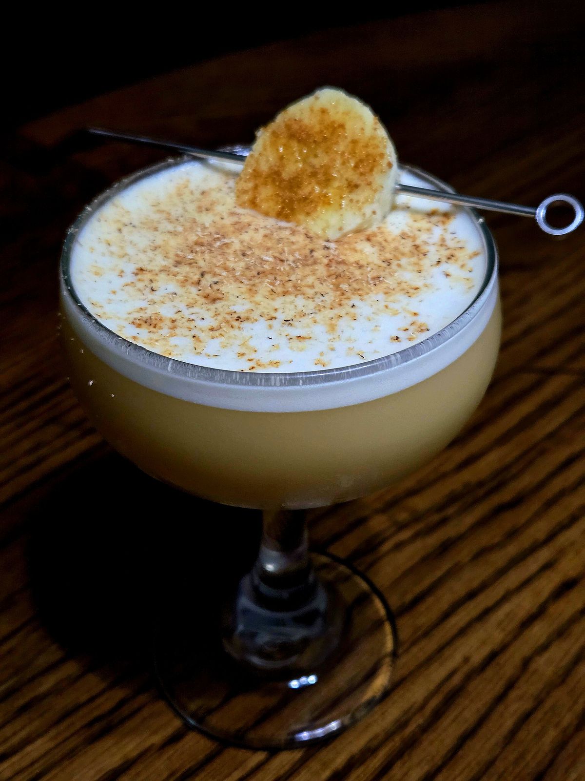 In Harm's ay flip with grated nutmeg and banana disc garnish with coconut sugar atop a wooden table.