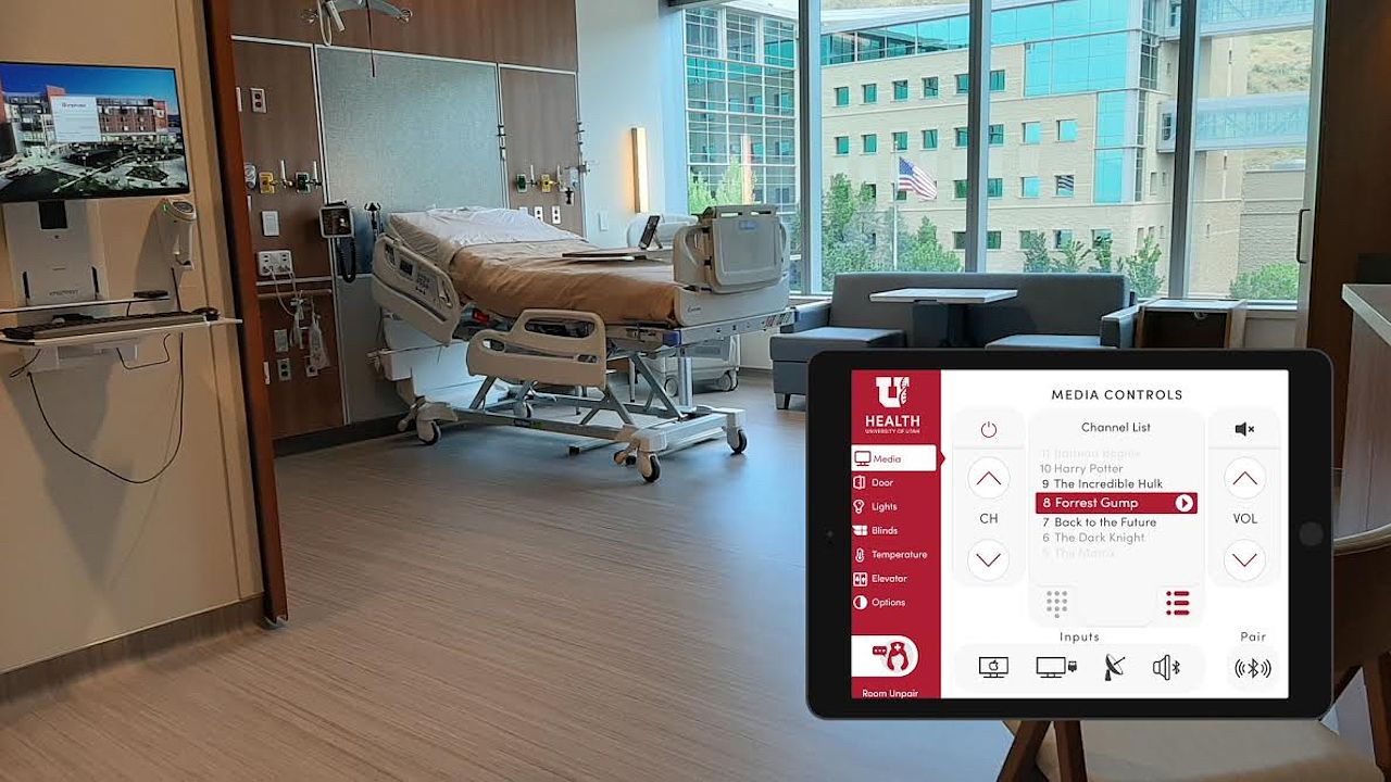 the inside of a hospital room showing a patient bed a large window,  a sofa, and lots of space. In the foreground of the frame is also an iPad that is running the custom smart room control app.